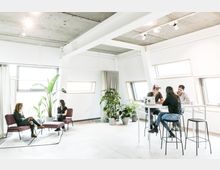 Heller, moderner Büroraum mit großen, schrägen Fenstern und Beton-Decke; im Vordergrund eine Sitzecke mit zwei Personen, daneben ein hoher Tisch mit drei Personen, umgeben von Zimmerpflanzen.