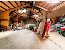 Innenansicht einer großen Scheune oder Lagerhalle mit hohem Satteldach, sichtbaren Holzbalken und Oberlichtern. Auf dem Betonboden stehen Werkzeuge, Regale, Bretter und verschiedene gelagerte Gegenstände entlang der Wände.