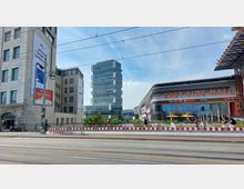 Straßenszene mit moderner Architektur: Links ein mehrstöckiges Gebäude aus hellem Stein mit Werbebannern, im Hintergrund ein hoher Glas-Büroturm, rechts das abgerundete, futuristisch gestaltete Einkaufszentrum "East Side Mall" mit leuchtendem Schriftzug. Im Vordergrund ein Gehweg mit Absperrungen und Straßenbahnschienen.
