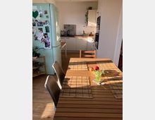 Ein moderner, heller Küchen- und Essbereich mit Holztisch, vier Stühlen und Bambus-Tischsets. Im Hintergrund eine helle Einbauküche mit Mosaik-Fliesenspiegel, sowie ein mintgrüner Kühlschrank mit angehefteten Fotos und Notizen.