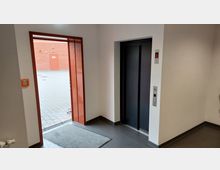 Ansicht eines Eingangsbereichs mit einer Aufzugstür auf der rechten Seite und einer offenen Glastür, die nach draußen führt. Draußen erkennbar ist eine gepflasterte Fläche und eine rote Backsteinwand mit einer verschlossenen Tür.