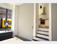 Ein Innenraum mit einer grauen Wand und einem großen Fenster, durch das eine gelbe Straßenbahn und ein Fußgänger draußen sichtbar sind. Rechts führt eine kurze Treppe in eine helle, minimalistische Küche mit weißen Schränken, einer Dunstabzugshaube und Hängelampe.