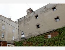 Außenansicht eines alten Gebäudes mit bröckelnder Fassade, teils freiliegenden Ziegelsteinen und kleinen Fenstern, daneben ein mit Efeu bewachsenes Backsteingebäude. Eine große Straßenlampe hängt an einem Kabel vor hellem Himmel.