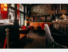 Innenraum einer stilvollen Bar mit freiliegender Backsteinwand, dunkler Holztheke mit Flaschenregal und warmen Leuchten. Vor großen Fenstern stehen kleine Tische mit roten Polstersesseln und einer gepolsterten Sitzbank entlang der Wand.