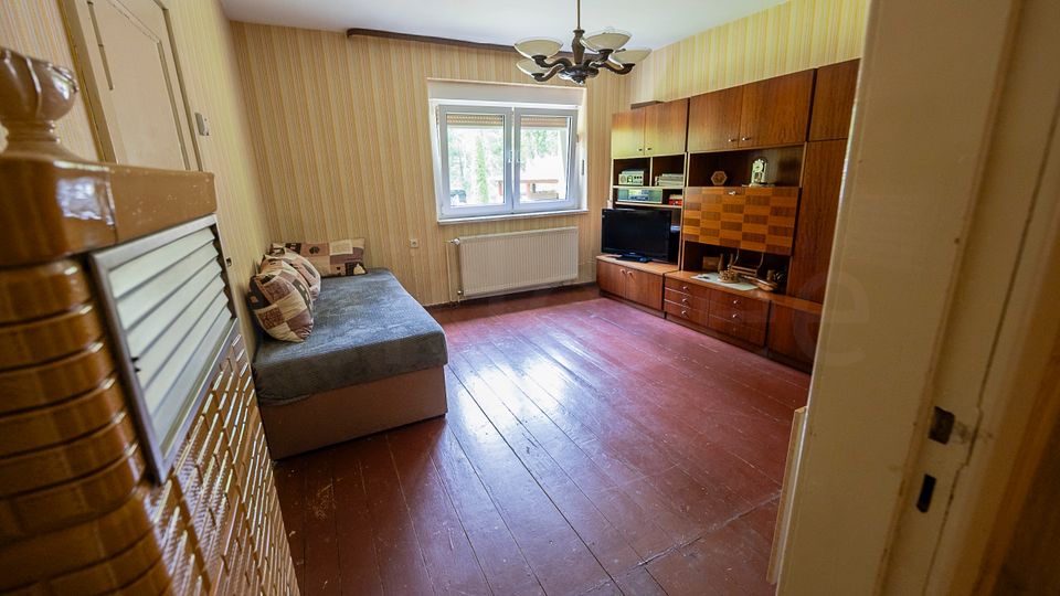 Schmales Wohnzimmer mit gestreifter Tapete, rotem Dielenboden und Deckenleuchte; an der Stirnseite ein Fenster mit Heizkörper darunter. Rechts steht eine große dunkle Wohnwand mit Fernseher, links ein Sofa, im Vordergrund ist der Eingang mit Kachelofenansicht zu sehen.