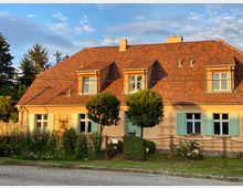 Ein Einfamilienhaus mit pastellgelber Fassade, türkisfarbenen Fensterläden und einem roten Ziegeldach. Vor dem Haus befindet sich ein Vorgarten mit blühenden Sträuchern, Blumen und einem kleinen Baum, umgeben von einem niedrigen Metallzaun.
