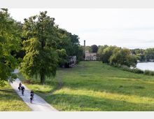 Weite Grünfläche mit Bäumen und einem geschotterten Weg, auf dem mehrere Personen mit Fahrrädern fahren. Im Hintergrund ein historisches Gebäude mit einem Schornstein, umgeben von Wald und in der Nähe eines Flusses.