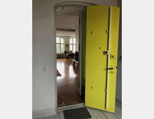 Blick durch eine offene, auffällig gelbe Tür in einen hellen Raum mit Holzboden, großen weißen Fenstern und einer Einrichtung aus Holztisch, Stühlen und Pflanzen. Im Vordergrund eine Türschwelle und ein grauer Teppich.