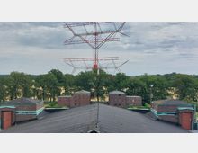 Backsteinbauten und ein Dach mit dunkler Eindeckung im Vordergrund, im Hintergrund eine große rot-weiße Stahlkonstruktion (Sendemast) und dahinter eine Landschaft mit dichten Bäumen unter bewölktem Himmel.