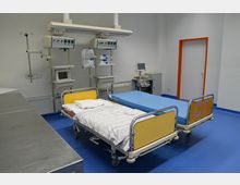 Krankenhauszimmer mit zwei Untersuchungsliegen, einer davon mit gelbem Kopfteil und der Aufschrift „CT“. An der Wand montiert sind medizinische Geräte, Regale und ein Monitor, der an eine Liege angeschlossen ist. Der blaue Boden und orangefarbene Türrahmen prägen den Raum.