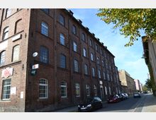 Backsteingebäude mit mehreren Stockwerken, großen Bogenfenstern und Werbeschildern an der Fassade, gelegen an einer ruhigen, von Autos gesäumten Straße mit Bäumen am rechten Straßenrand.