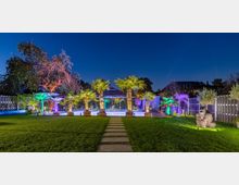 Gartenlandschaft bei Nacht mit beleuchtetem Swimmingpool, umgeben von Palmen und bunten Lichteffekten. Im Vordergrund ein gepflegter Rasen mit Gehwegplatten, rechts eine große Gorilla-Statue, im Hintergrund ein Haus und Hecken.