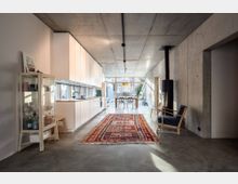 Modernes Wohnzimmer mit offener Küche, Sichtbetonwänden und glänzendem Betonboden. Eine lange Küchenzeile mit weißen Schränken erstreckt sich entlang einer Seite, ein bunter Teppich bedeckt den Boden, und ein Holzofen sowie große Fensterfronten im Hintergrund sorgen für eine helle, minimalistische Atmosphäre.