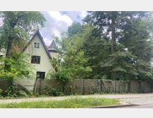 Wohnhaus mit spitzem Giebel und einem kleinen Turm, umgeben von dichten Bäumen und einem begrünten Garten. Im Vordergrund ist ein Drahtzaun mit Rankpflanzen und eine Gehwegfläche zu sehen.