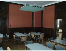 Ein Innenraum mit roten Wänden und dunkler Holzvertäfelung, ausgestattet mit mehreren Tischen und schlichten Stühlen in Holz- und Metalloptik. Eine moderne, rechteckige Deckenleuchte hängt zentral über dem Raum, und durch eine geöffnete Tür ist ein angrenzender Bereich sichtbar.