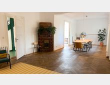 Wohnzimmer mit Holzboden und einer offenen Tür, die zu einem grünen Treppenbereich führt. Im Raum stehen ein brauner Kachelofen mit Pflanzen darauf, ein Esstisch mit mehreren Stühlen und ein Sessel, sowie große Topfpflanzen und eine Hängelampe.