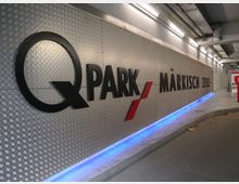 Eine Innenansicht eines Parkhauses mit einer silbernen Wand aus Riffelblech, auf der der Schriftzug „Q-Park Märkisch Zeile“ in schwarzen und roten Buchstaben angebracht ist. Der Boden ist gebogen, bläuliche Beleuchtung verläuft entlang der Wand, und die Decke verfügt über sichtbare Rohrleitungen.