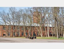 Backsteingebäude mit großen Fenstern, umgeben von kahlen Bäumen im Vordergrund. Auf dem Gelände stehen mehrere Räder und Achsen, vermutlich aus Metall, auf Paletten.