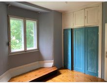 Kleiner Raum mit Holzdielenboden, einem Fenster mit Blick auf grüne Bäume und einer Einbauschrankwand mit weißen und türkisfarbenen Türen. Die Wände sind grau gestrichen, und ein niedriges Podest befindet sich unterhalb des Fensters.