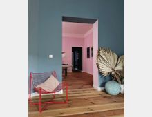 Wohnraum mit Holzboden und Wänden in Blau und Rosa. Im Vordergrund ein roter Drahtsessel mit einem Kissen, dekorative getrocknete Palmenblätter in Vasen und eine pastellblaue Kugelvase; im Hintergrund ein Essbereich mit Lampen und Wandbildern.