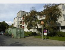 Mehrstöckiges Wohngebäude mit hellgrauer Fassade und Balkonen, umgeben von Bäumen und einer Wiese. Im Vordergrund stehen ein Baustellencontainer sowie ein Halteverbotszeichen, während rechts daneben ein gepflasterter Gehweg und eine Straße sichtbar sind.