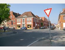 Straßenszene in einer Kleinstadt mit Backsteinfassaden, darunter ein zweistöckiges Gebäude mit Schaufenstern und einem Geschäft. Mehrere geparkte Autos stehen entlang der Straße, ein Vorfahrt-gewähren-Schild ist im Vordergrund zu sehen, und ein Baum steht links.