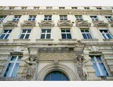 Fassade eines historischen Gebäudes mit weißem Putz und mehreren Stockwerken, großen rechteckigen Fenstern sowie dekorativen Elementen wie Statuen von Figuren, die einen Balkon stützen, und einer verzierten Arkade über einem Eingang.