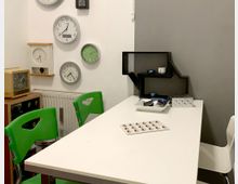 Kleiner Raum mit weißem Tisch und bunten Elementen, an dessen Wand mehrere analoge Uhren in unterschiedlichen Designs hängen. Daneben steht ein Regal in Sprechblasenform, während grüne und weiße Stühle um den Tisch platziert sind.