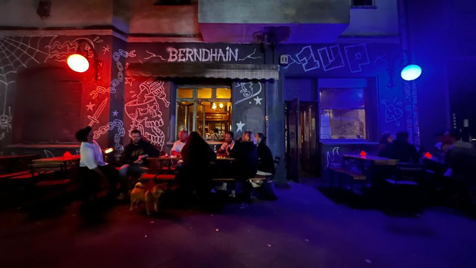 Straßenansicht eines Eingangs zu einer Bar namens „BERNDHAIN“ mit beschmierter Fassade und Graffiti, beleuchtet von roten und blauen Außenlampen. Vor dem Eingang stehen mehrere Holztische mit Bänken auf dem Gehweg, dahinter sind Fenster und ein kleiner Balkon an der Gebäudefront zu sehen.