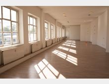 Langer, leerer Innenraum mit weißen Wänden, hellem Holz- bzw. Laminatboden und einer Reihe großer Sprossenfenster entlang der linken Seite. Unter den Fenstern stehen Heizkörper, und das Tageslicht wirft rechteckige Schattenmuster auf den Boden.