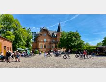 Historisches Backsteingebäude mit spitzem Turm umgeben von Bäumen und einer gepflasterten Platzfläche, auf der sich Menschen und Fahrräder befinden; im Hintergrund ein kleines Café mit Sonnenschirmen und ein Imbisswagen rechts.