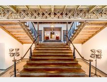 Holztreppe in einem historischen Gebäude mit reich verzierten Geländern und Holzbalkendecke, flankiert von zwei dekorativen Stehlampen. Im oberen Bereich des Raumes befinden sich Sitzmöbel und warme Beleuchtungselemente.