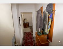 Schmaler Flur mit hellem Anstrich und rotem Teppichläufer, rechts ein Holzspiegel mit Hut, Mantel, Regenschirm und Telefon auf einem Schränkchen; im Hintergrund ein Wohnbereich mit Kommode, Tischlampe und Spiegel.