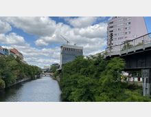 Ein städtischer Kanal mit dichtem Grün an den Ufern, flankiert von modernen Gebäuden, darunter ein hohes Glasgebäude mit Baukränen. Im Vordergrund verläuft eine erhöhte Brücke aus Metall rechts im Bild.