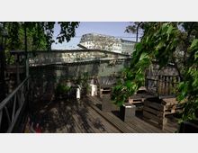 Dachterrasse mit Holzdeck und Sitzmöbeln aus Paletten, umgeben von Pflanzen und Netzen. Im Hintergrund sind moderne Gebäude und Bäume zu sehen.