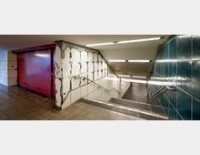 Unterführung mit Treppen, Wänden aus weißen und türkisfarbenen Fliesen, teilweise beschädigten Wänden mit freiliegendem Mauerwerk und Graffiti. Der Bereich ist mit rechteckigen Deckenleuchten beleuchtet, und Geländer verlaufen entlang der Treppen.