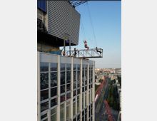 Ein Hochhaus mit einer verglasten Fassade, auf dessen Dach eine kleine Plattform mit einer hängenden Schaukel montiert ist, auf der mehrere Personen zu sehen sind. Im Hintergrund erstreckt sich eine Stadtlandschaft mit Straßen und Gebäuden.