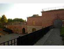 Historisches Festungsgebäude aus rotem Backstein mit Rundbogenfenstern und Eingangstoren, umgeben von gepflasterten Wegen und einem schmiedeeisernen Geländer. Im Hintergrund befinden sich Bäume sowie eine erhöhte, begrünte Fläche.