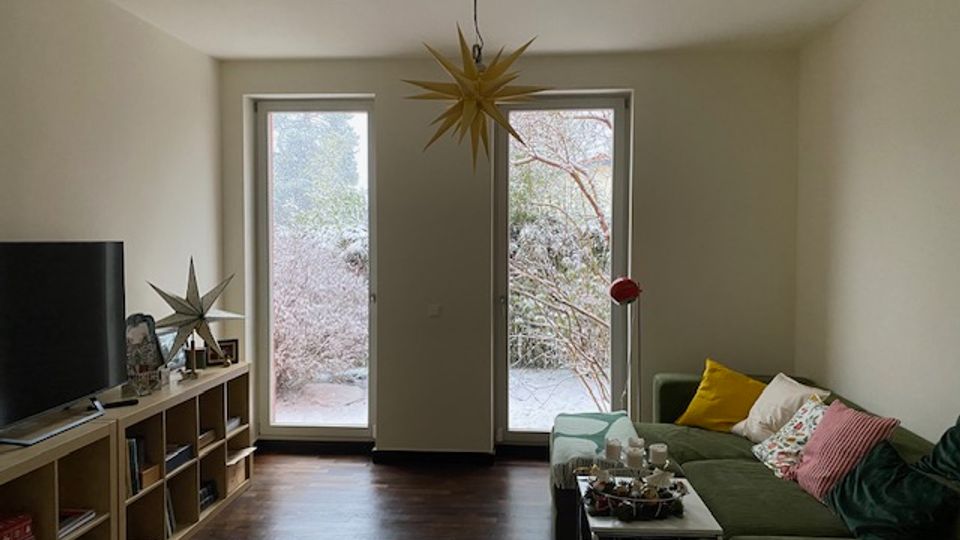 Wohnzimmer mit dunklem Holzboden, grünem Ecksofa und Couchtisch, links ein Fernseher auf einem niedrigen Regal. Zwei bodentiefe Fenster/Terrassentüren geben den Blick in einen Garten frei, an der Decke hängt eine sternförmige Pendelleuchte.