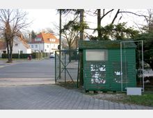 Grüne Holzhütte mit abblätternder Farbe und einem kleinen Fenster, daneben ein Maschendrahtzaun mit offenem Tor; im Hintergrund stehen Wohnhäuser mit roten Dächern entlang einer ruhigen Straße, wo ein Fahrradfahrer vorbeifährt.
