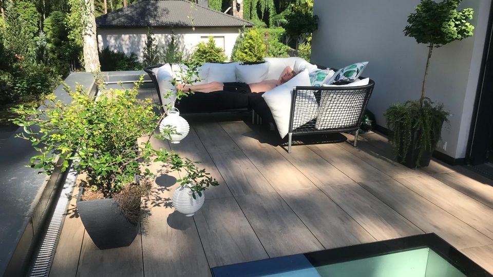 Moderne Dachterrasse mit Holzdeck, schwarzen Loungemöbeln und weißen Kissen sowie mehreren Pflanzkübeln und hängenden Laternen. Im Hintergrund sind ein flaches Nebengebäude und dichter Wald zu sehen; am Terrassenrand befindet sich ein verglaster Bodenausschnitt.