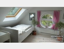 Dachzimmer mit einem Bett und einer Kommode, Tageslicht durch ein großes Dachfenster und ein rundes Fenster mit rosa Vorhängen. Zimmerpflanzen und helle Holzböden ergänzen den schlicht eingerichteten Raum.