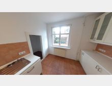 Kleine Küche mit weiß gestrichenen Wänden, einer Arbeitsfläche mit Spülbecken, weißen Schränken und terrakottafarbenen Fliesen auf dem Boden. Ein großes Fenster mit Blick auf einen Außenbereich und ein Heizkörper befinden sich an der Wand.