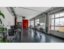 Großraumbüro mit hohen weißen Wänden, großen Fenstern mit Jalousien und schwarzem Boden. Es gibt mehrere Schreibtische mit Bürostühlen, Computern und Pflanzen, sowie einen roten Vorhang im Hintergrund.