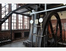 Innenraum einer historischen Industrieanlage mit großen Zahnrädern und zwei Druckanzeigen, umgeben von Backsteinwänden und hohen Fenstern mit Metallrahmen. Eine Metalltreppe führt zu einer höheren Ebene, und im Hintergrund sind historische Fotografien an der Wand ausgestellt.