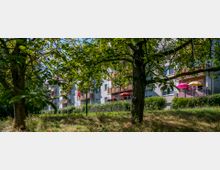 Mehrstöckiges Wohngebäude mit Balkonen, teilweise mit bunten Sonnenschirmen, umgeben von Grünflächen und Bäumen im Vordergrund. Die Szene zeigt eine ruhige Wohngegend an einem sonnigen Tag.