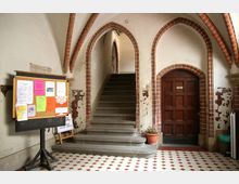 Eingangsbereich eines historischen Gebäudes mit gemustertem Fliesenboden, gotischen Spitzbögen aus Backstein, einer Steintreppe, einer Holztür und einer Pinnwand mit farbigen Aushängen. Eine Topfpflanze steht neben der Treppe, die Wände zeigen Abnutzungsspuren.