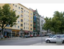 Straßenszene mit mehrstöckigen Wohn- und Geschäftsgebäuden, darunter ein gelbes und ein grünes Haus. Im Erdgeschoss befinden sich Geschäfte wie ein Asia-Imbiss und eine Fahrschule. Parkende Autos und ein Gehweg mit Bäumen sind im Vordergrund sichtbar.