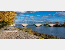 Steinbrücke über einen Fluss mit mehreren Bögen, dahinter Bäume und ein bewölkter Himmel. Im Vordergrund ein gepflasterter Uferbereich mit einem Geländer und herbstlichem Laub.