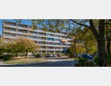 Wohnblock in einer Wohnsiedlung mit fünf Etagen und Balkonen, teilweise mit bunten Markisen und Blumenkästen. Im Vordergrund ein Parkplatz mit geparkten Autos, umgeben von Bäumen und herbstlichem Laub.